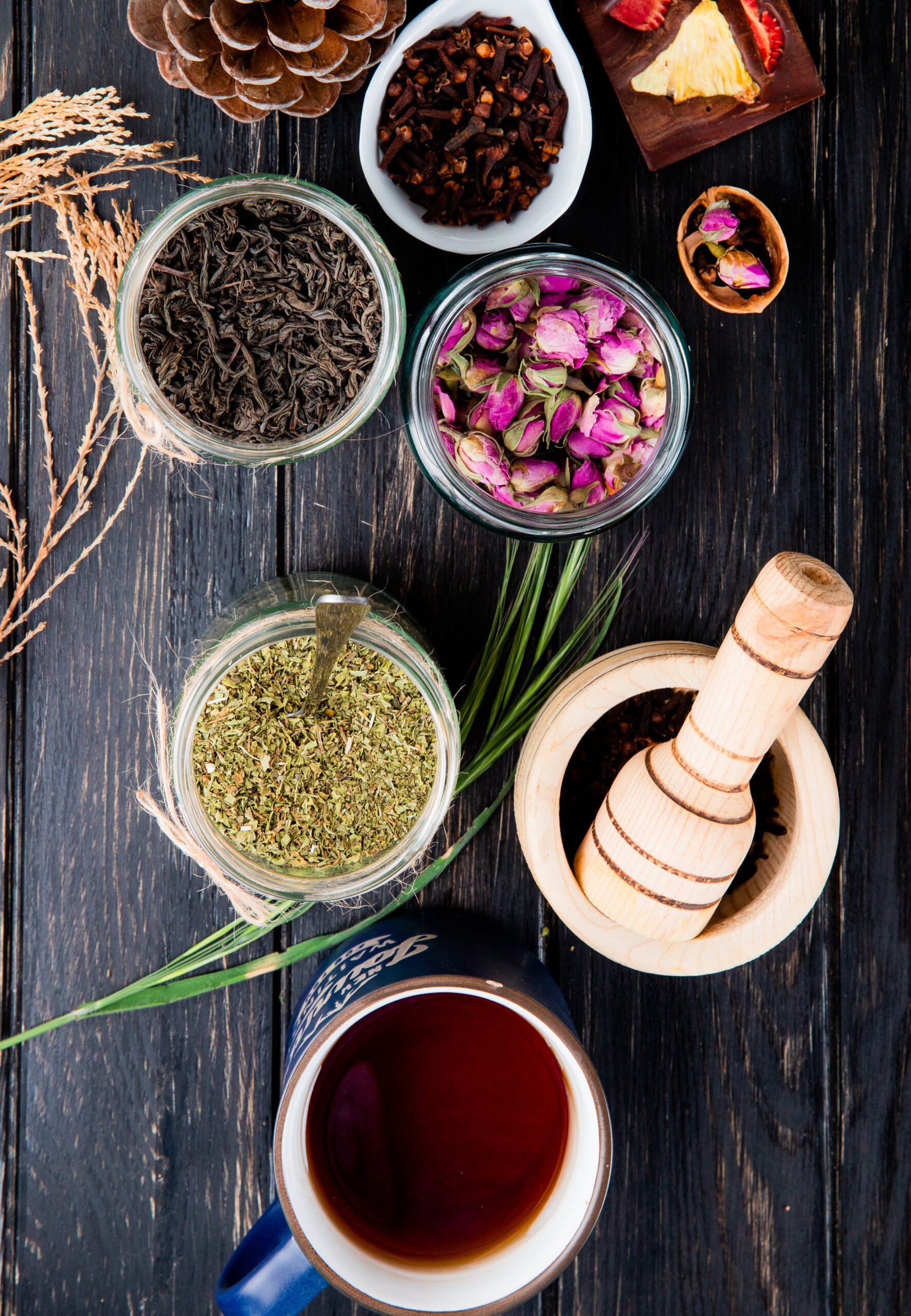 top view of various spices and herbs dry black tea leaves, peppermint, rose buds, clove spice and black peppercorns in glass jars on black wooden background herbal medication